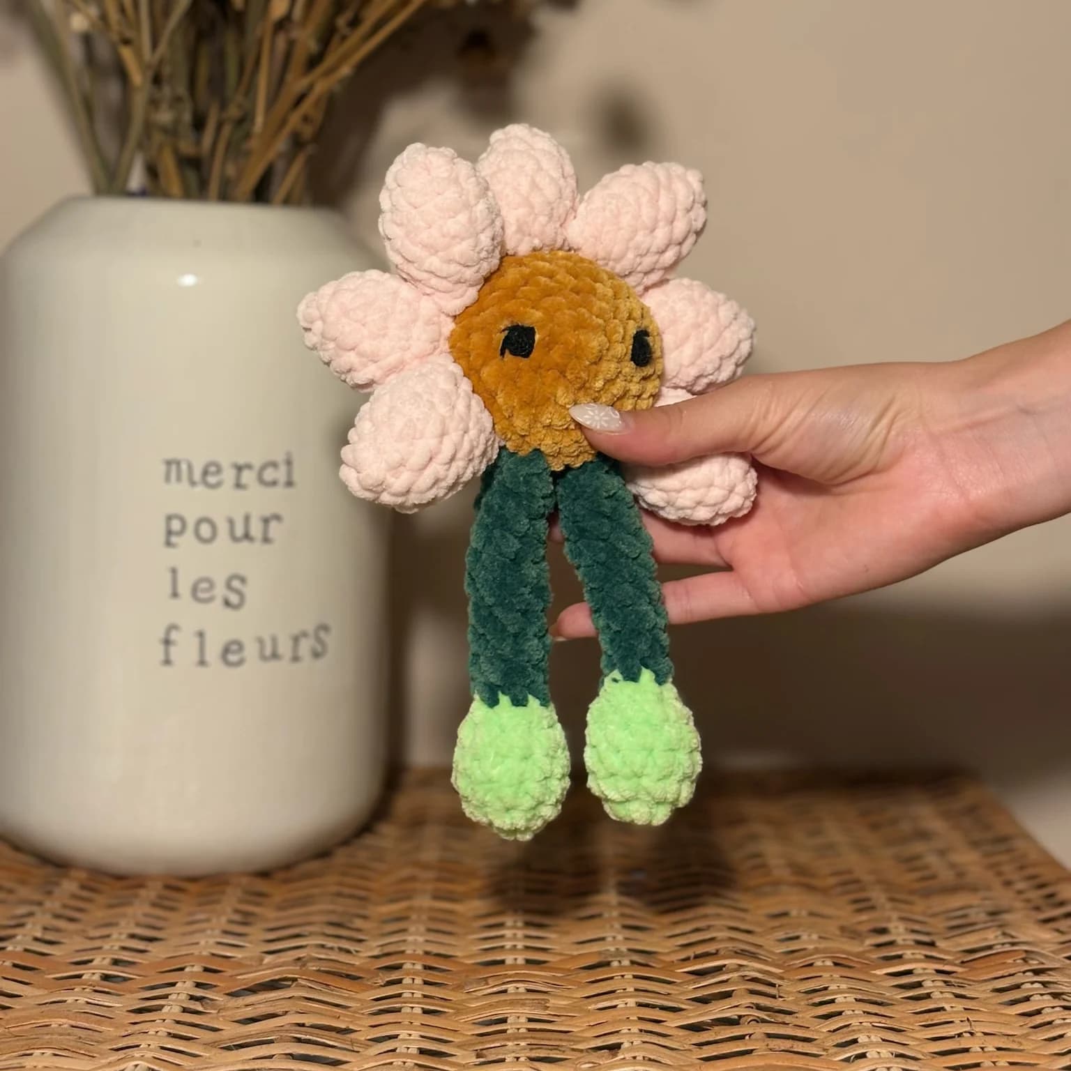 Peluche en laine en forme de fleur aux pétales roses, cœur marron avec yeux et longues jambes vertes.