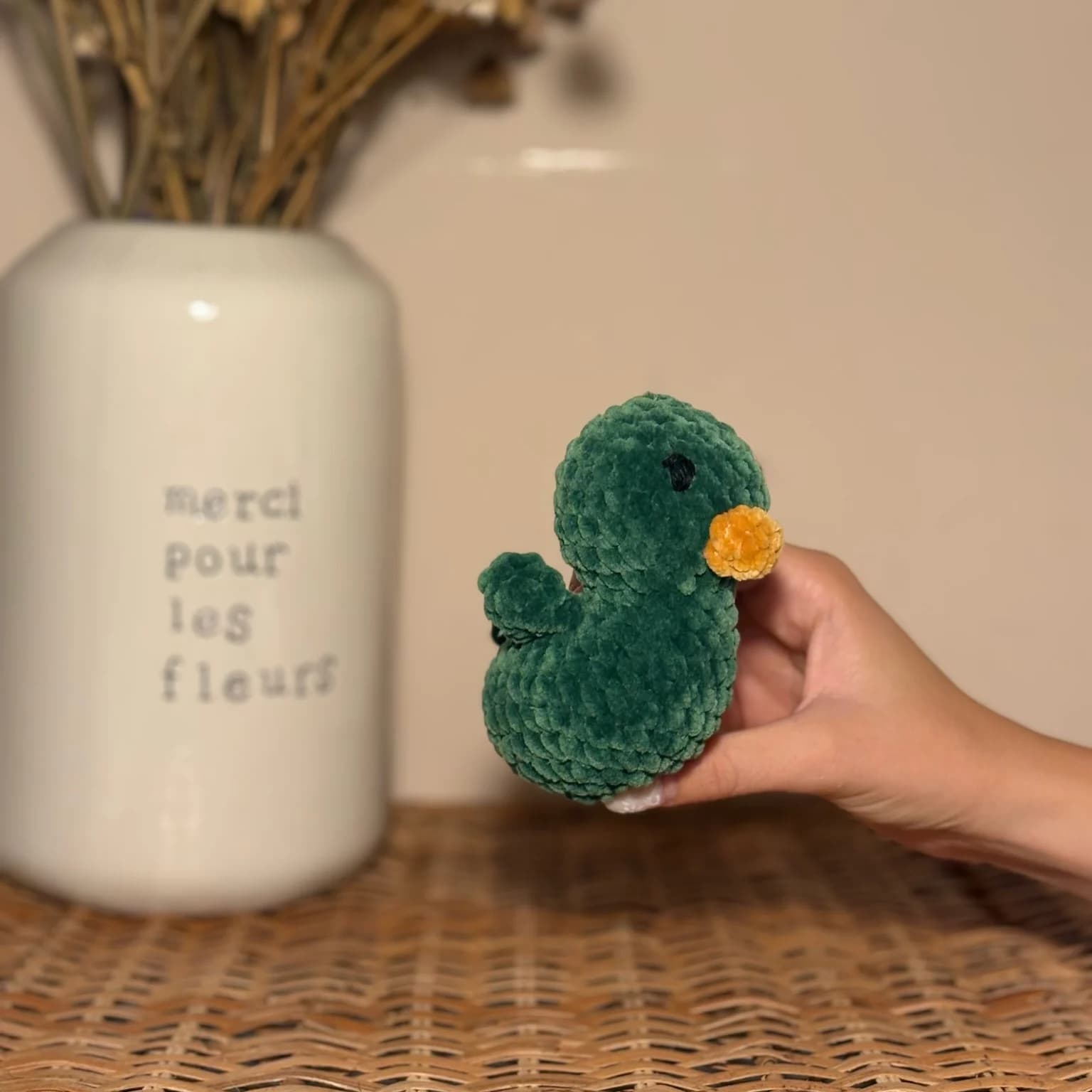 Main tenant une petite peluche de canard vert foncé au bec orange devant un vase blanc