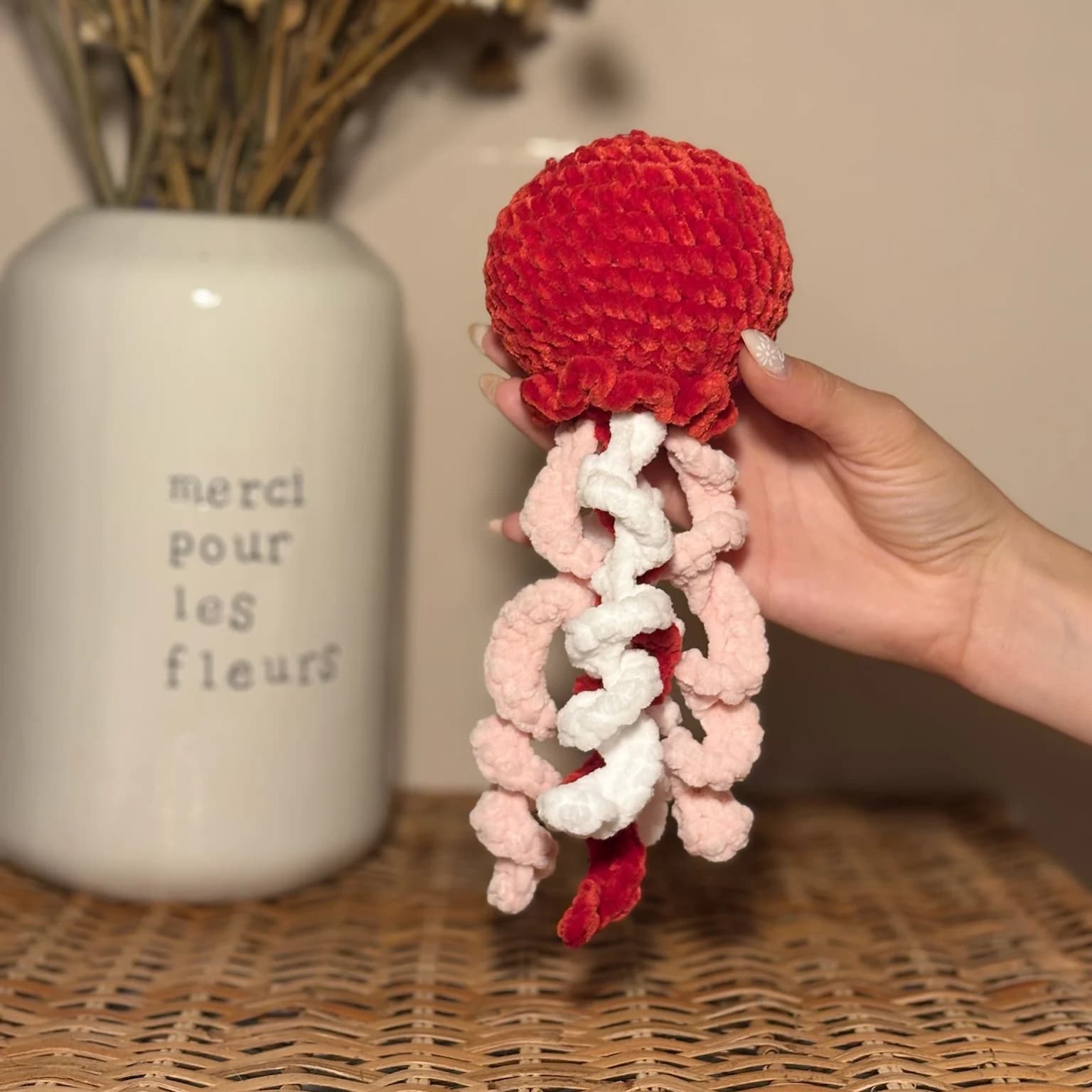 Peluche méduse en laine rouge à tentacules roses et blanches, tenue à la main devant un vase blanc.