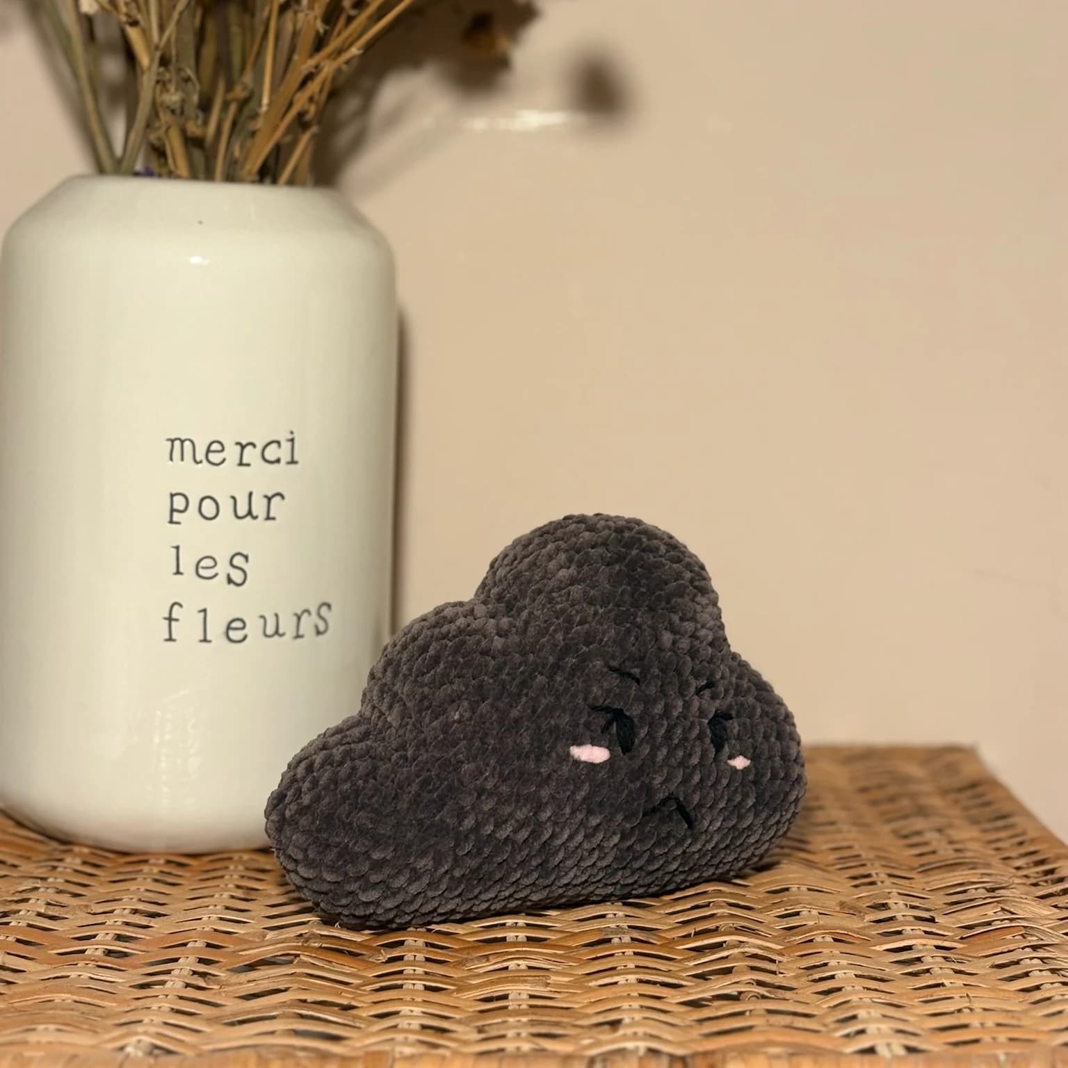 Peluche en forme de nuage gris foncé au visage brodé boudeur, posée sur un meuble en osier.