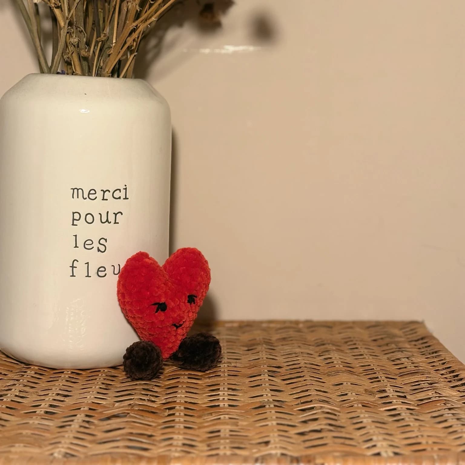 Cœur rouge en peluche avec visage souriant brodé et petits pieds foncés posé près d'un vase blanc
