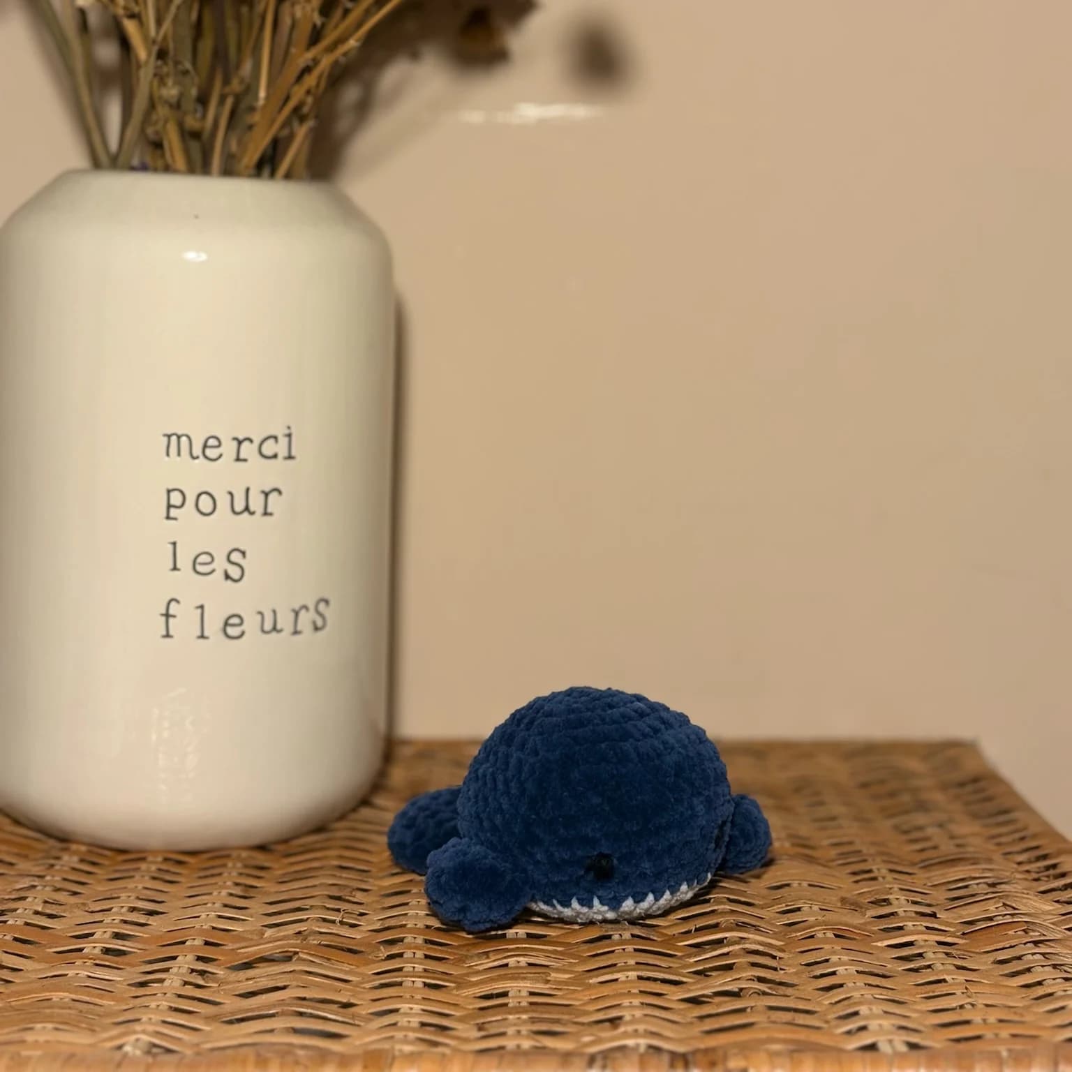 Peluche baleine bleu foncé posée sur un meuble en osier tressé près d'un vase blanc