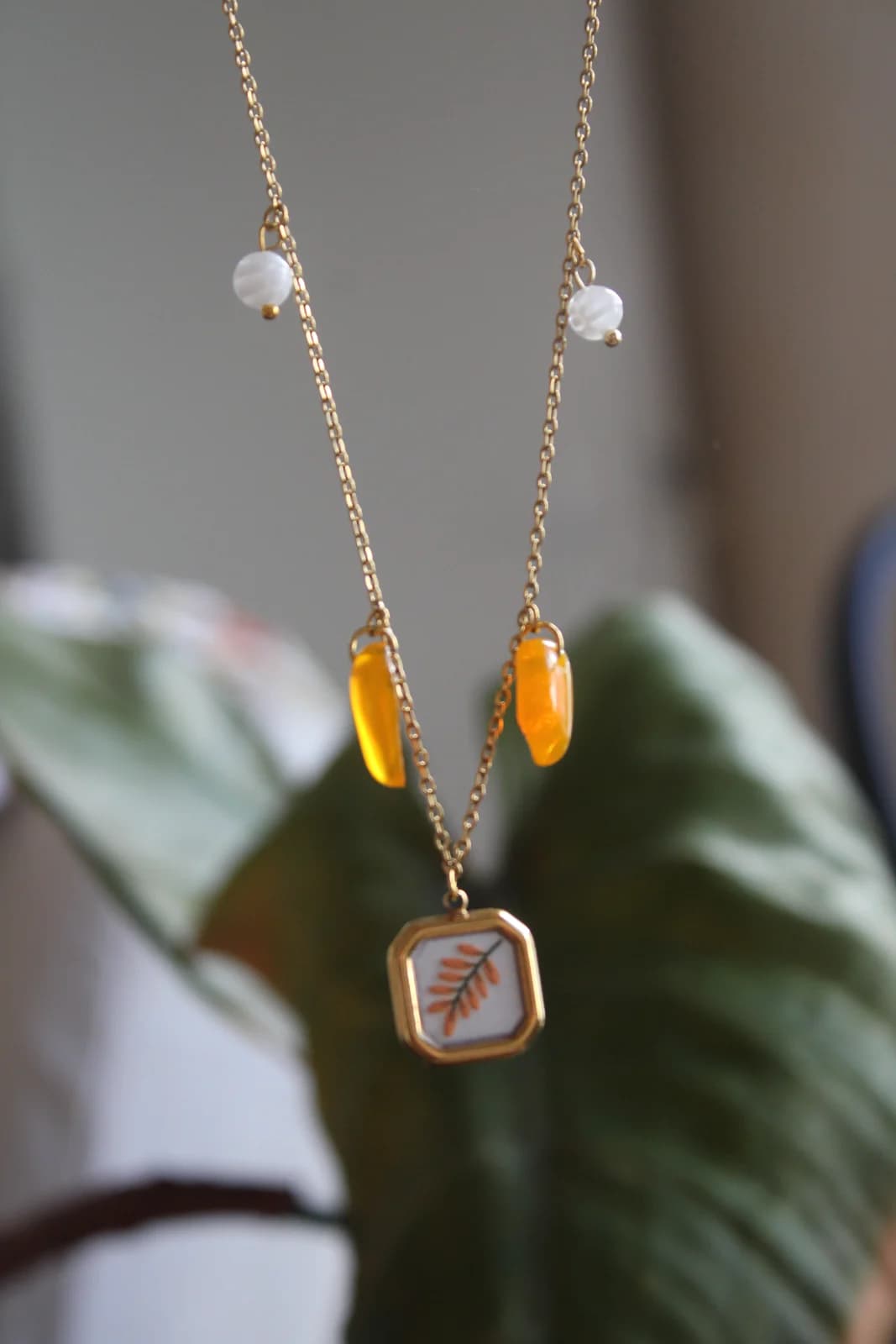 Détail du collier doré suspendu avec pampilles orange, perles blanches et pendentif feuillu.