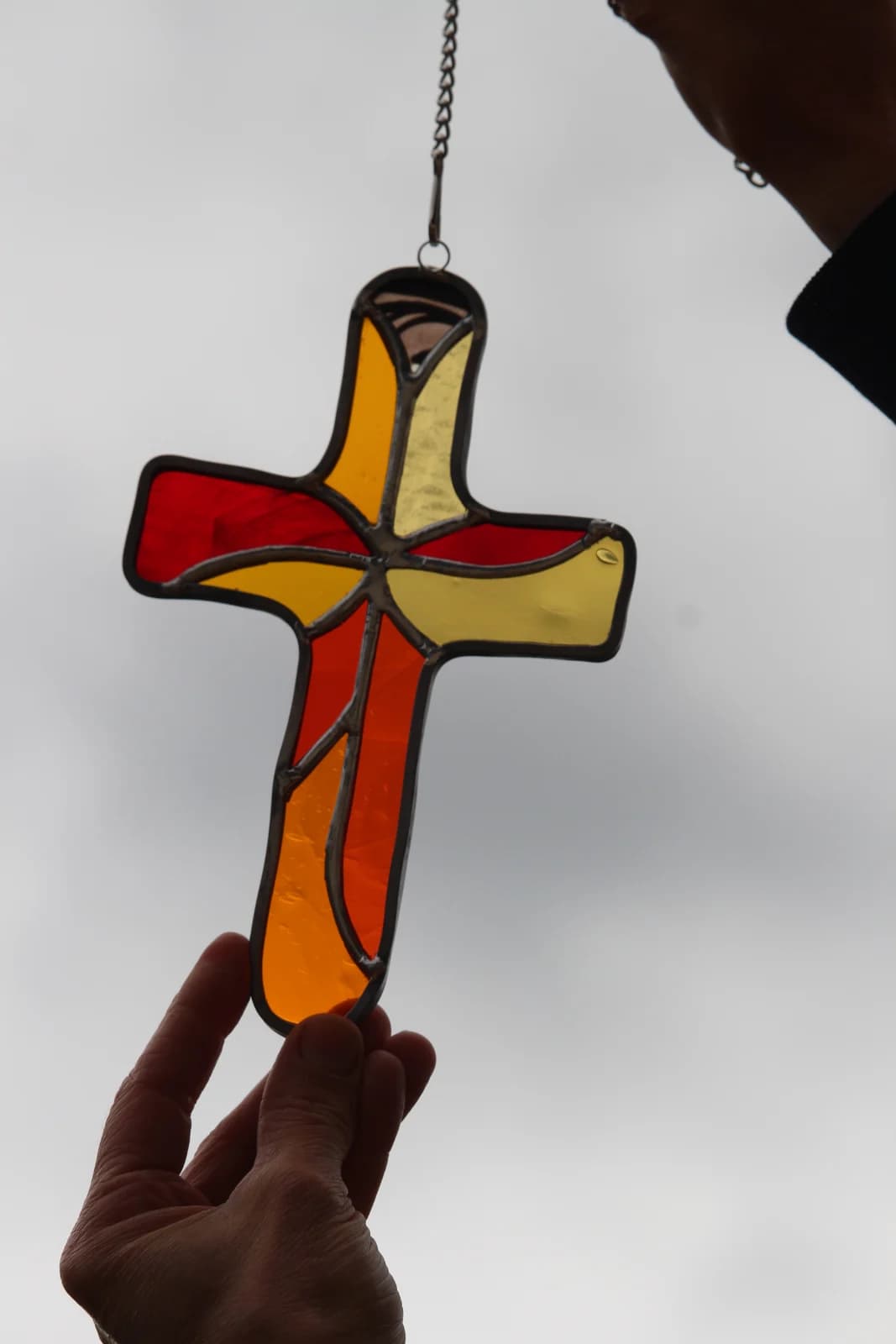 Croix à suspendre en vitrail tiffany aux nuances rouge, jaune et orange, tenue en extérieur devant l'herbe.