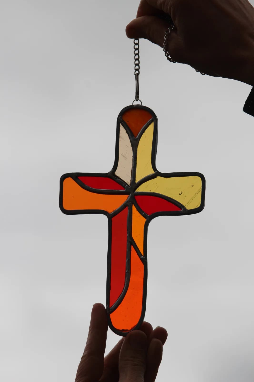 Croix en vitrail aux teintes orangées, rouges et jaunes tenue à la main en contre-jour