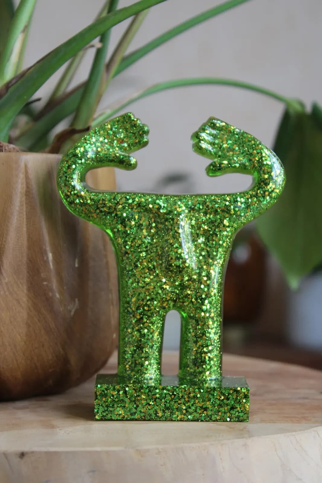 Statuette stylisée verte pailletée aux bras levés devant une plante d'intérieur.