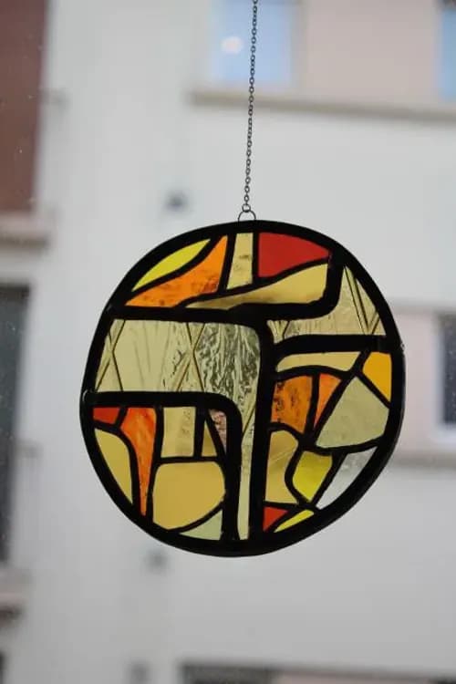 Vitrail rond abstrait aux tons chauds suspendu par une fine chaîne devant une fenêtre lumineuse.