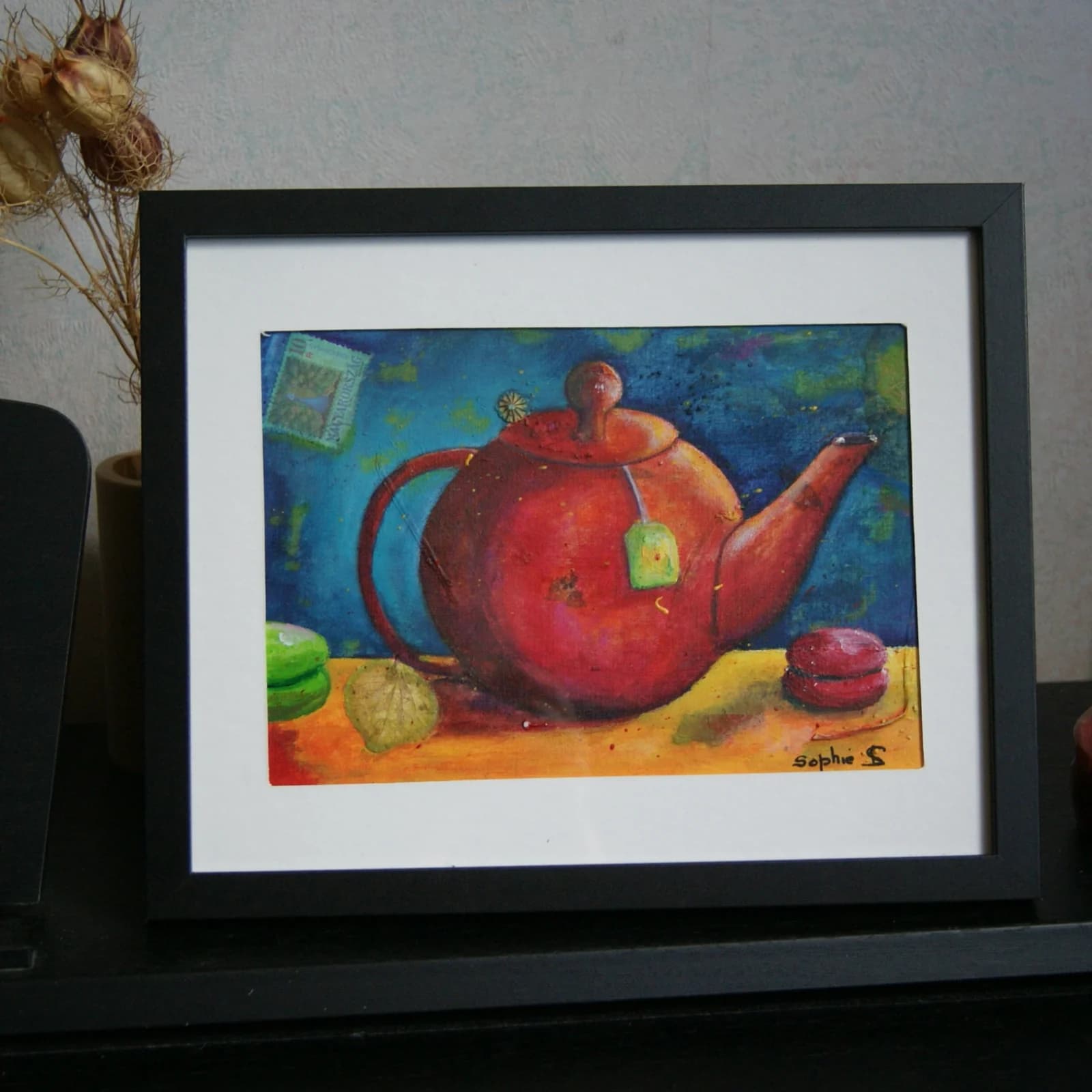 Tableau d'une théière rouge et de macarons sur fond bleu, présenté dans un cadre noir avec passe-partout.