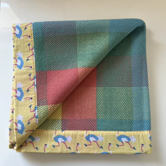 Plaid plié tissé à carreaux bleus, verts et roses avec bordure jaune ornée d'oiseaux.