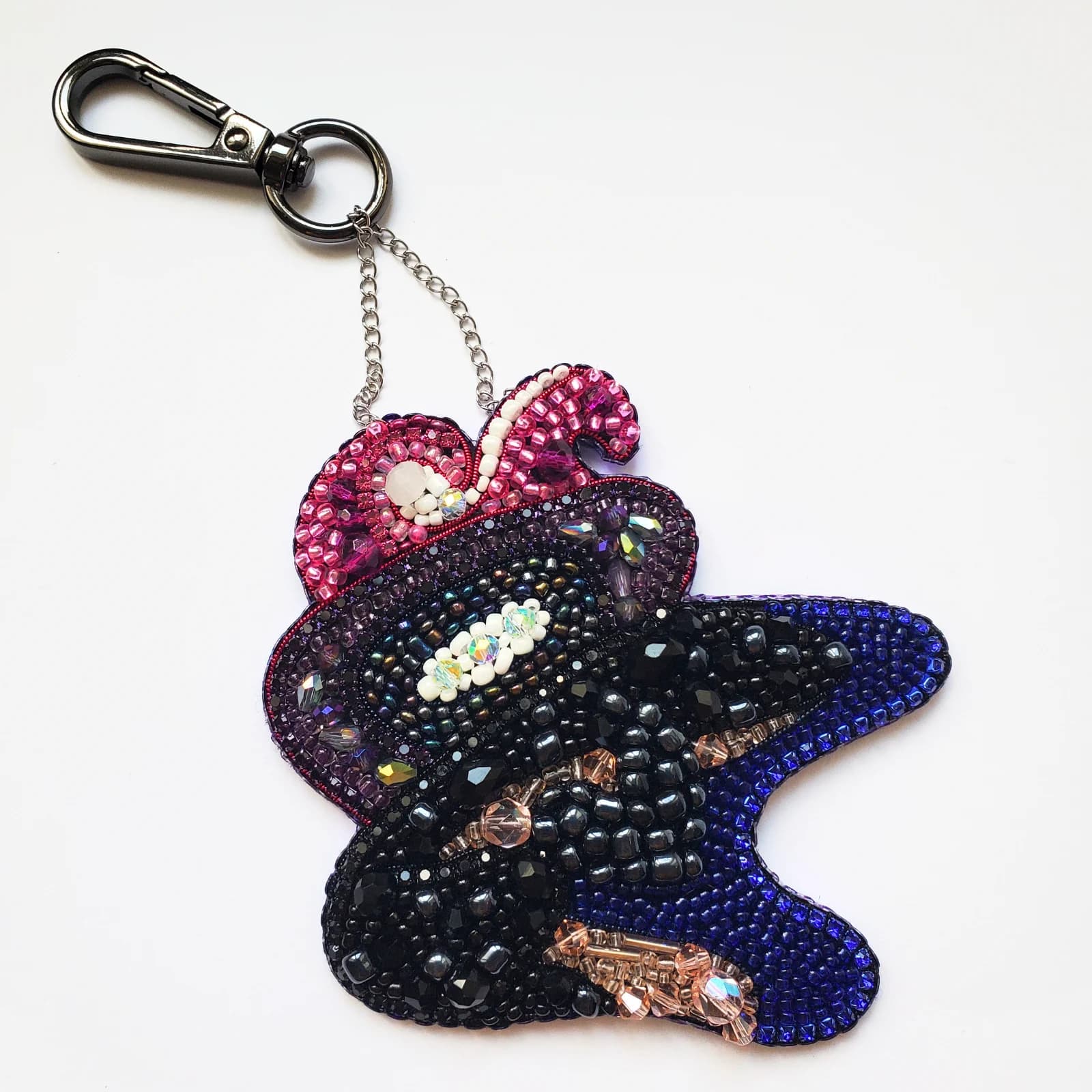 Porte-clé Hélios brodé de perles et sequins noirs, violets et roses avec mousqueton sur fond blanc.