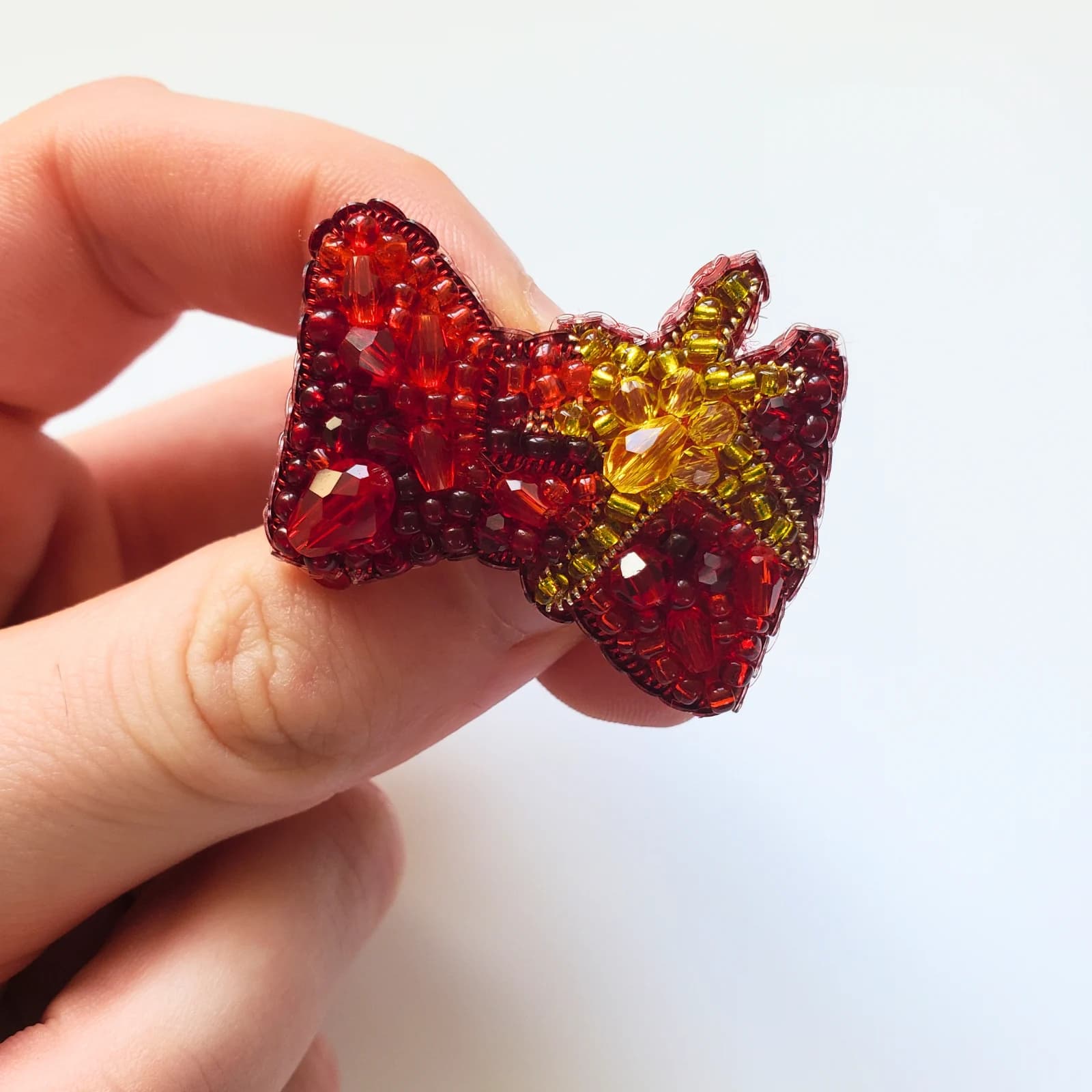 Broche en forme de nœud rouge et étoile dorée brodée de perles, tenue entre les doigts sur fond blanc.