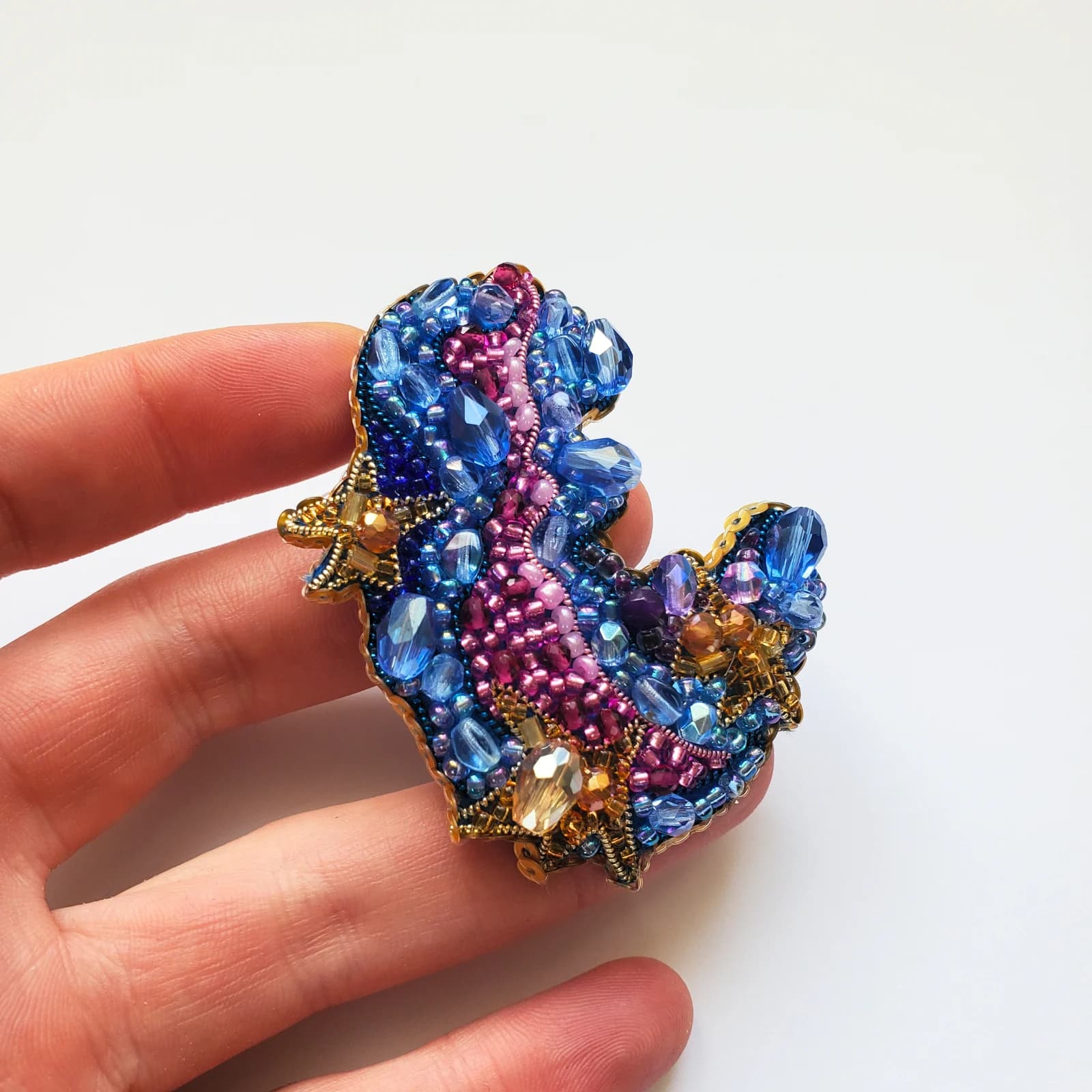 Broche perlée artisanale Nebula aux nuances de bleu, rose et doré tenue dans la main