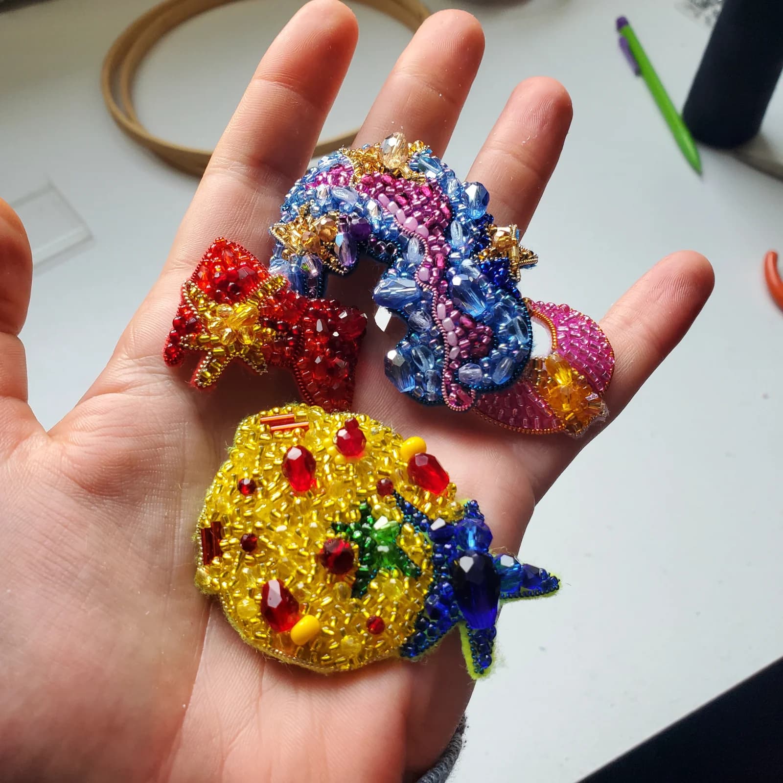 Trois broches artisanales brodées de perles colorées et sequins, présentées dans la paume d'une main.