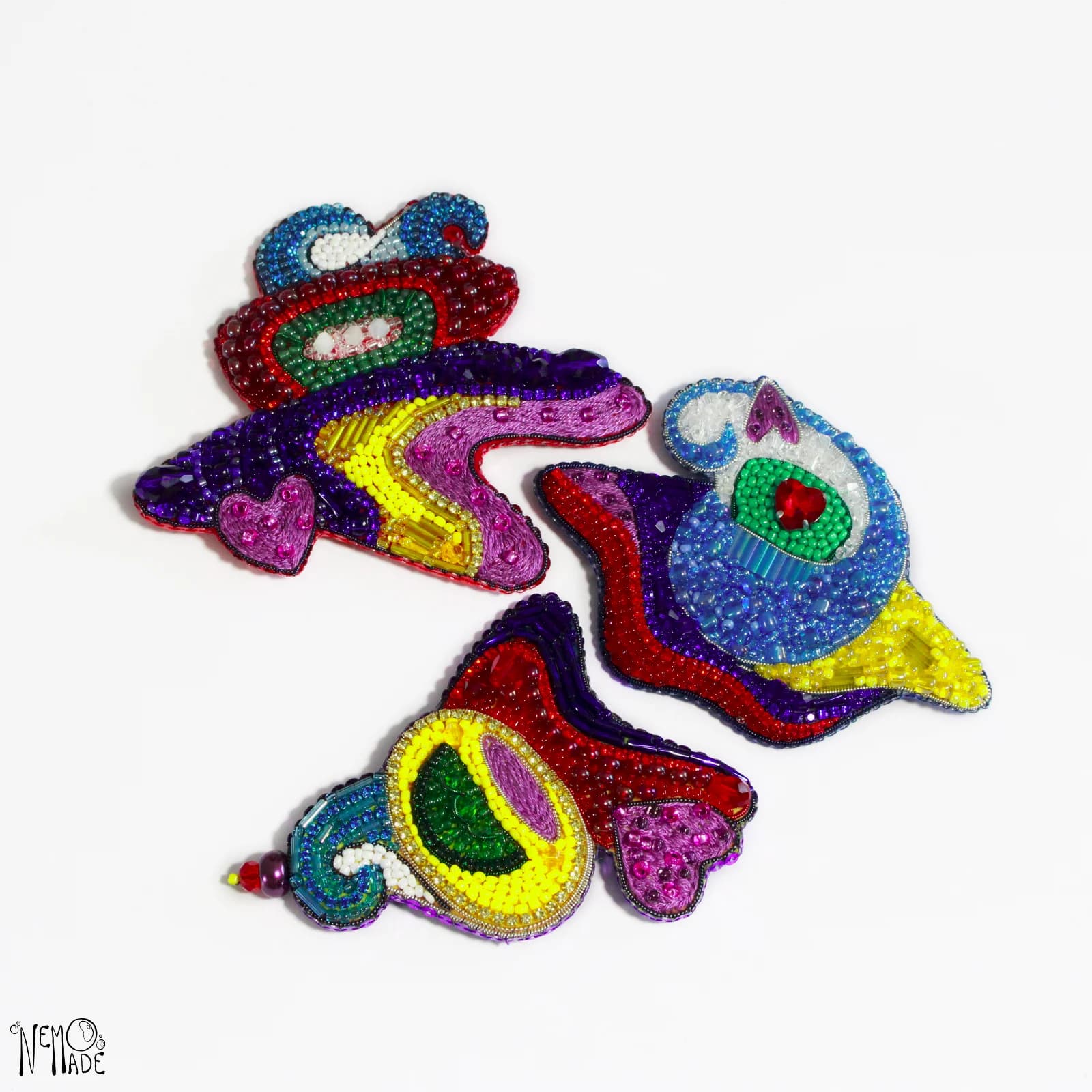Lot de trois broches artisanales aux formes abstraites brodées de perles multicolores vives sur fond blanc