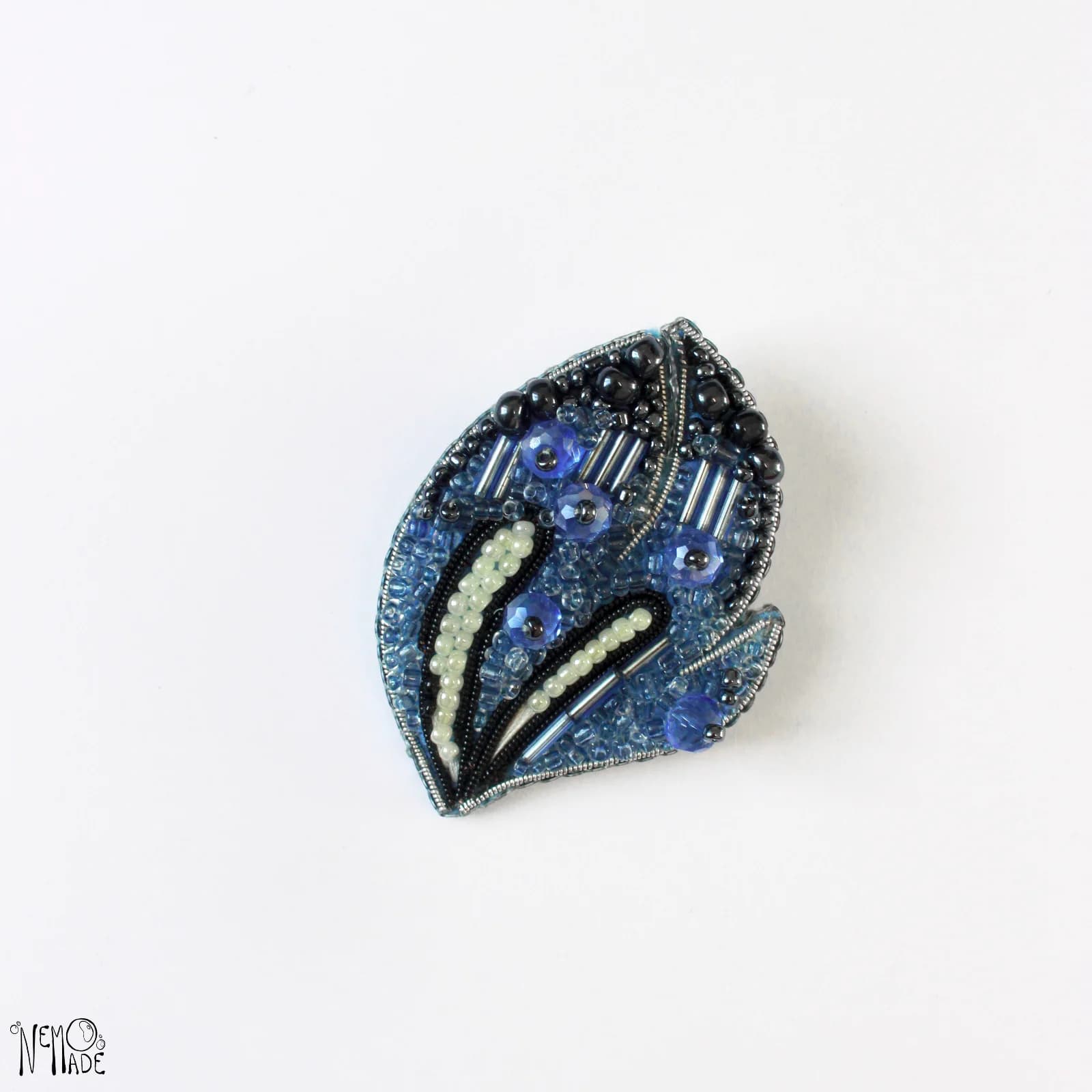 Broche brodée Lite Flame en perles et cannetille bleues, noires et claires sur fond blanc.