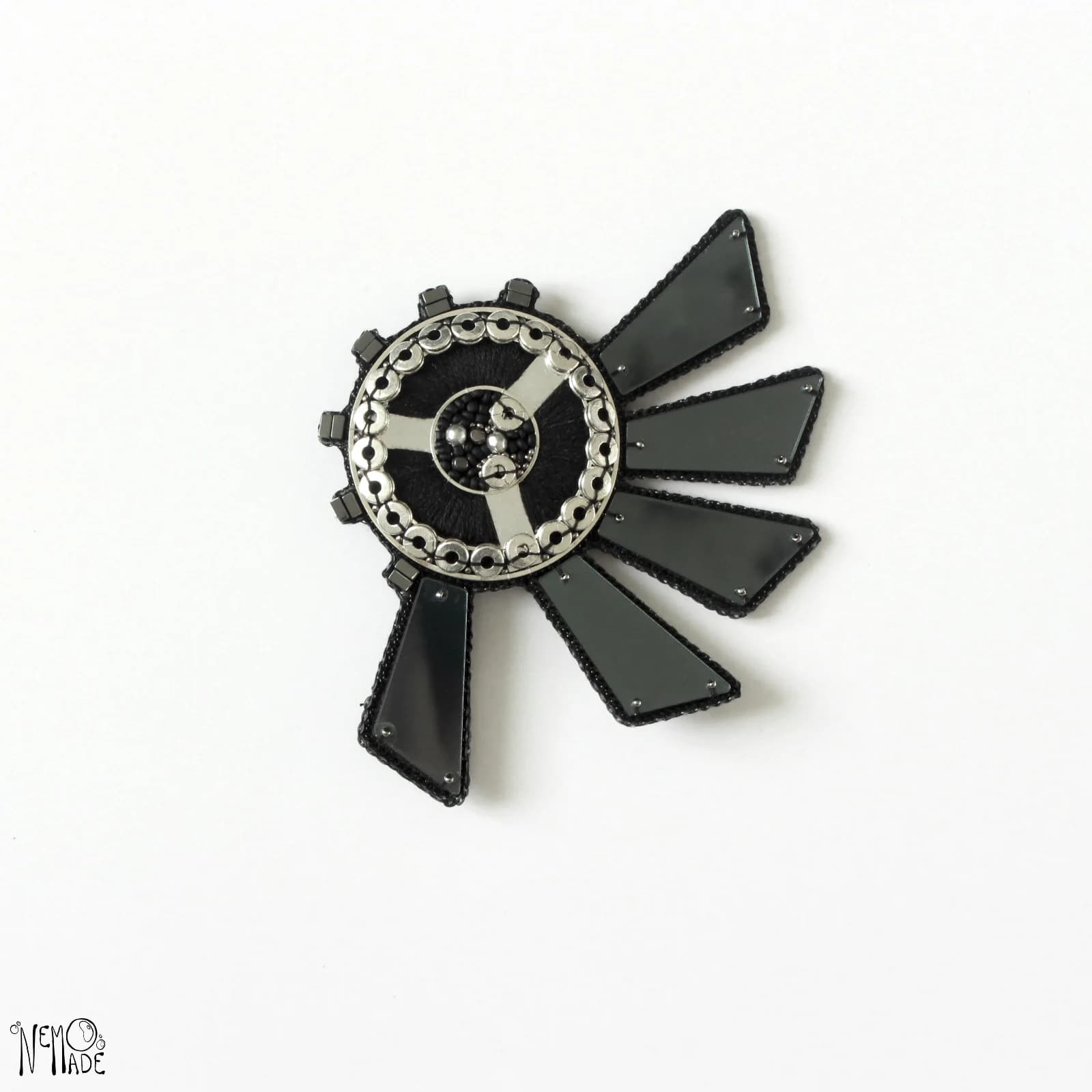 Broche Réflection graphique noire et argentée aux détails perlés sur fond blanc.