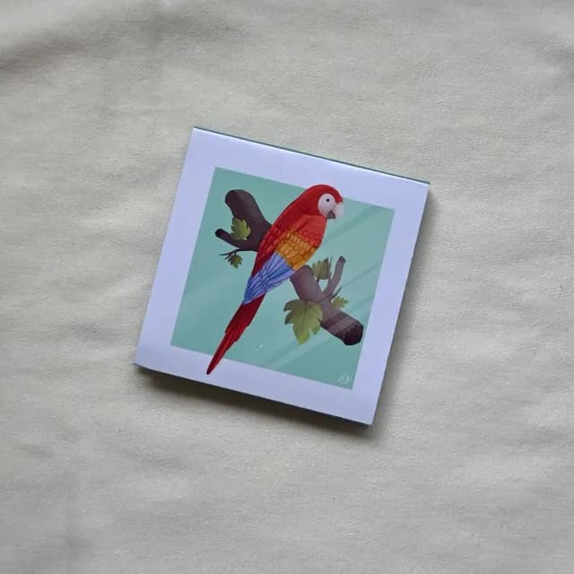 Bloc-notes carré orné d'un perroquet rouge et bleu perché sur une branche, posé sur tissu beige.