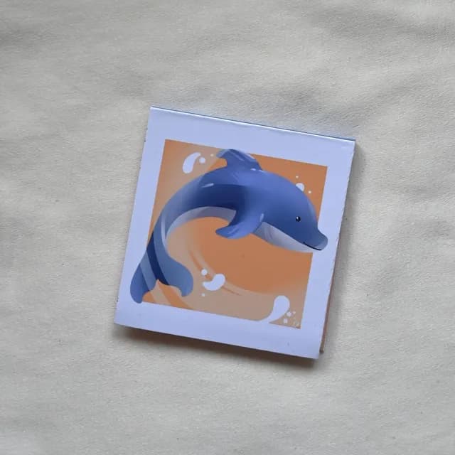 Bloc-notes carré illustré d'un dauphin bleu sautant sur un fond orangé et blanc.