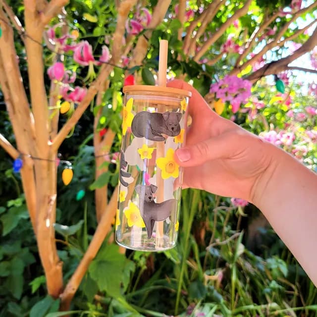Verre transparent tenu en main avec motifs labrador noir et fleurs jaunes, couvercle et paille en bambou.