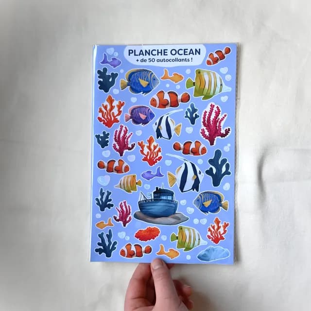 Planche d'autocollants thème océan tenue en main avec poissons multicolores, coraux et bateau.