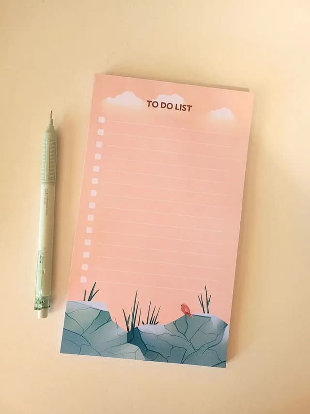 Bloc to-do list rose à lignes avec dessin de rochers et oiseau, posé à côté d'un porte-mine.