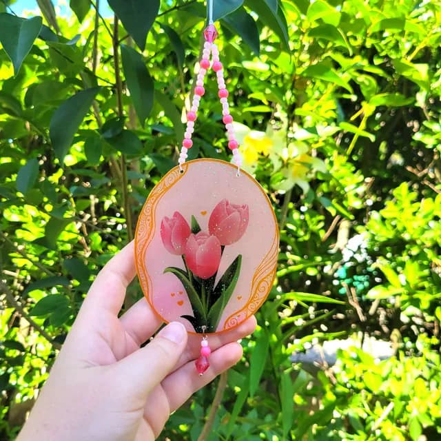 Suncatcher ovale tulipes roses tenu en main devant du feuillage, orné de perles roses suspendues
