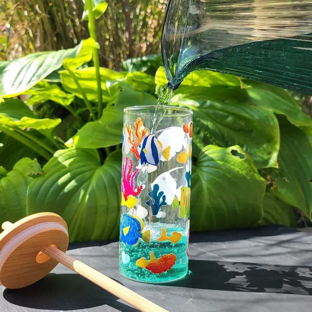 Eau versée dans le verre décoré de poissons et coraux, couvercle et paille en bambou posés à côté.