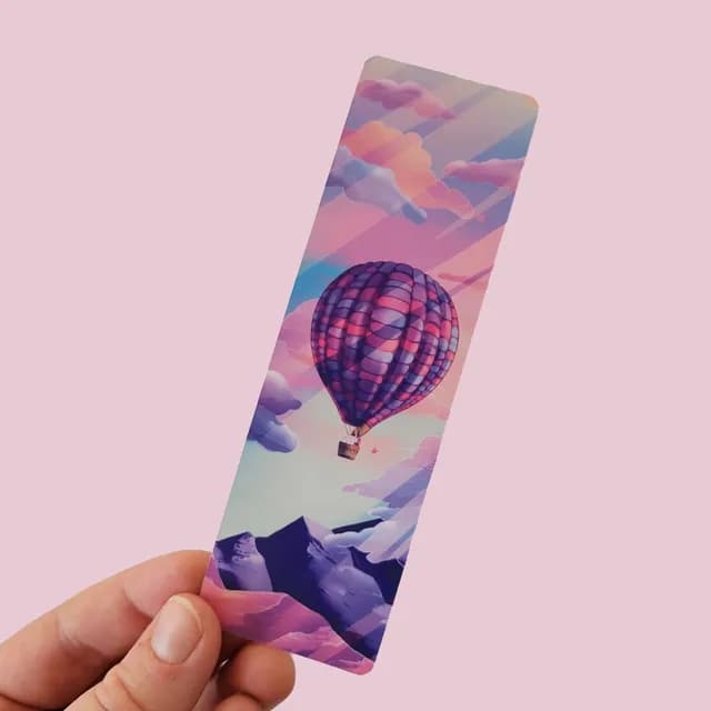 Marque-page tenu en main illustrant une montgolfière violette au-dessus de montagnes et nuages pastel.