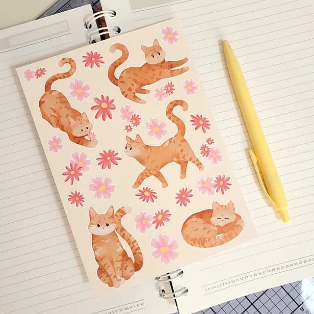 Planche de stickers avec chats roux et fleurs roses posée sur un carnet ouvert à côté d'un stylo jaune.