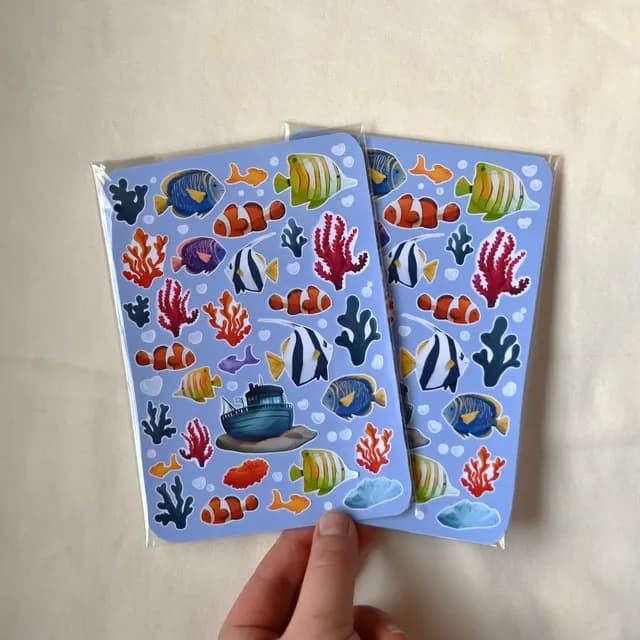 Deux planches de stickers sur le thème de l'océan avec poissons tropicaux et coraux, tenues en main.