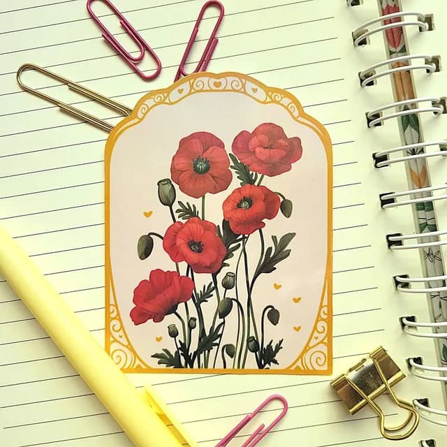 Sticker arche illustré de coquelicots rouges et bordure dorée, posé sur un carnet à spirales
