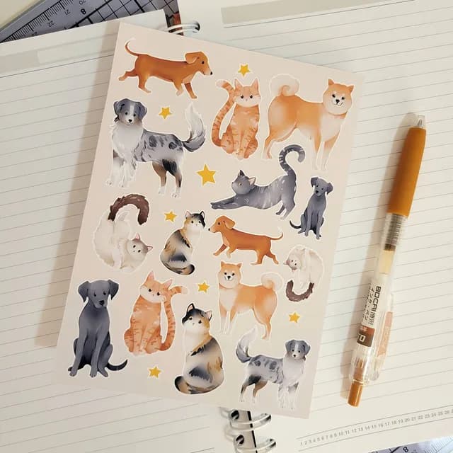 Planche d'autocollants illustrant des chiens et des chats aux tons roux et gris, posée sur un carnet à spirales.
