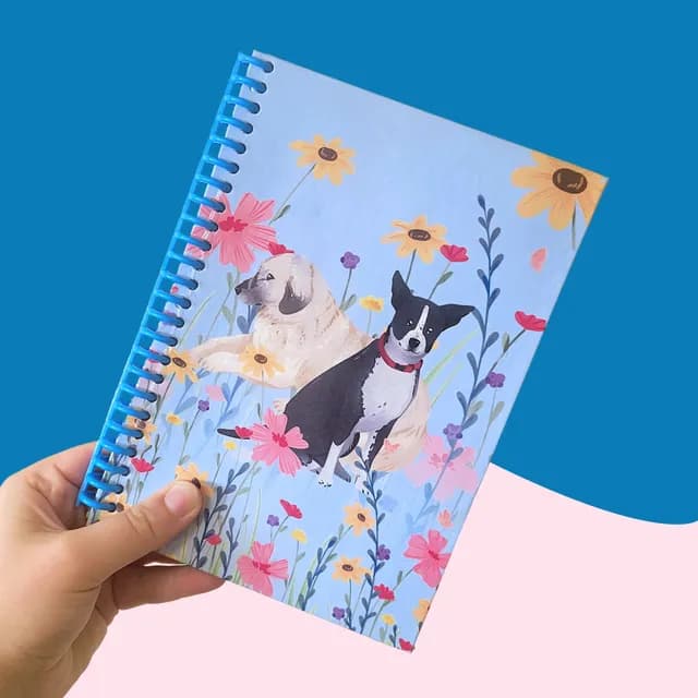 Carnet A5 à spirale bleue tenu en main, illustré de deux chiens parmi des fleurs colorées sur fond bleu ciel