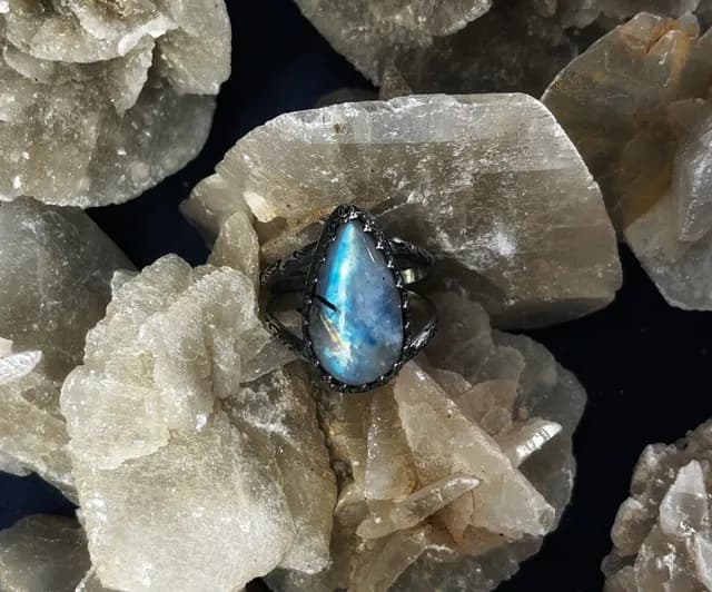 Bague en argent sertie d'une pierre de lune en goutte aux reflets bleus, posée sur une roche cristalline.