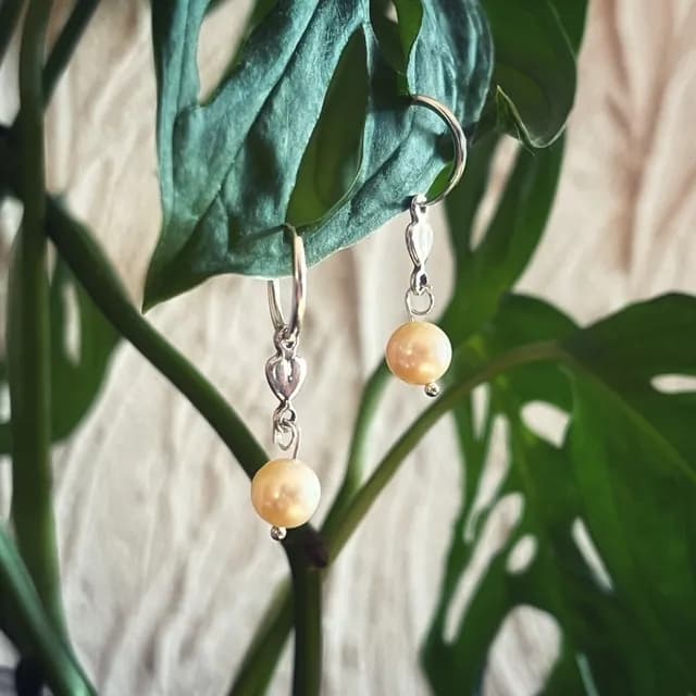 Créoles argentées ornées d'un motif cœur et d'une perle nacrée, suspendues à une feuille verte.