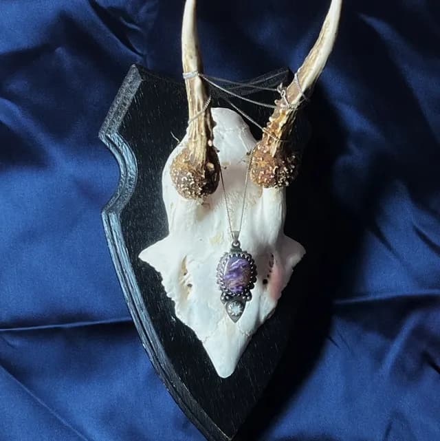 Pendentif Éclipse en argent avec charoite violette et opale, suspendu à un trophée de cornes sur fond bleu nuit.