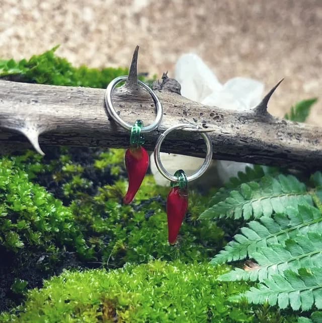Créoles argentées avec pendentifs cornicello en verre rouge et vert, posées sur une branche dans la mousse