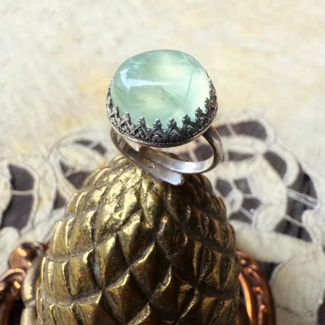 Bague en argent sertie d'un cabochon vert clair, posée sur un support métallique doré façon pomme de pin