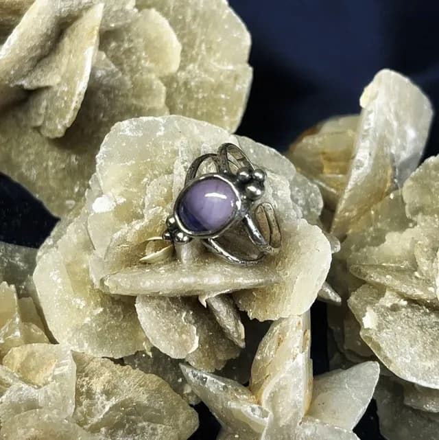 Bague en argent sertie d'un cabochon violet aux petites billes métalliques, posée sur une rose des sables.