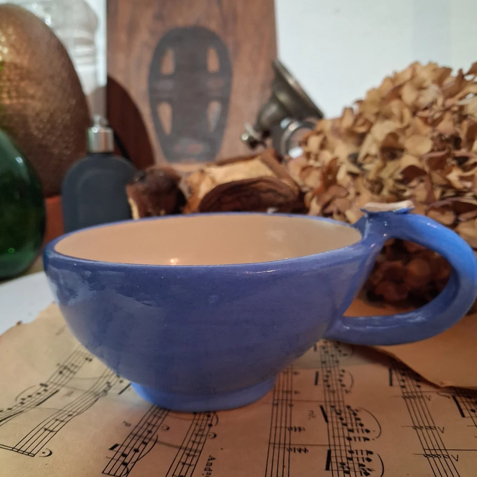 Tasse évasée en céramique bleue avec anse ornée d'un livre miniature, posée sur une partition de musique.
