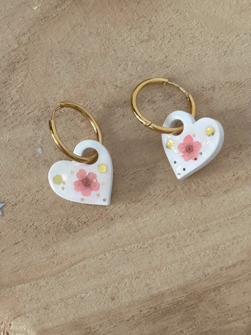 Anneaux dorés avec pendentifs cœurs blancs ornés d'une fleur rose et de paillettes dorées posés sur du bois.