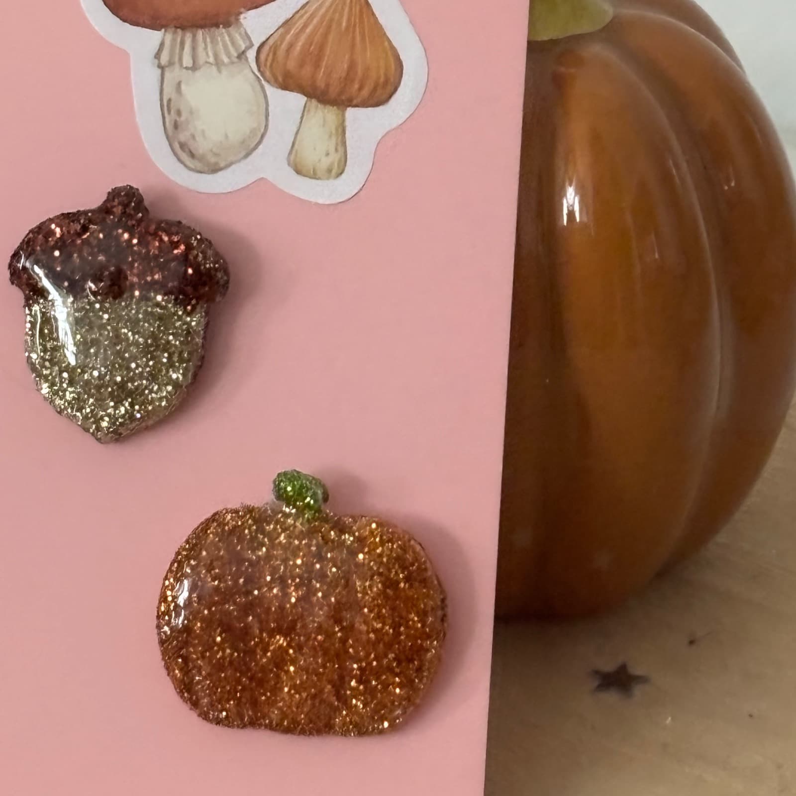 Pin's scintillants en forme de gland et de citrouille sur carte rose à côté d'une courge décorative.