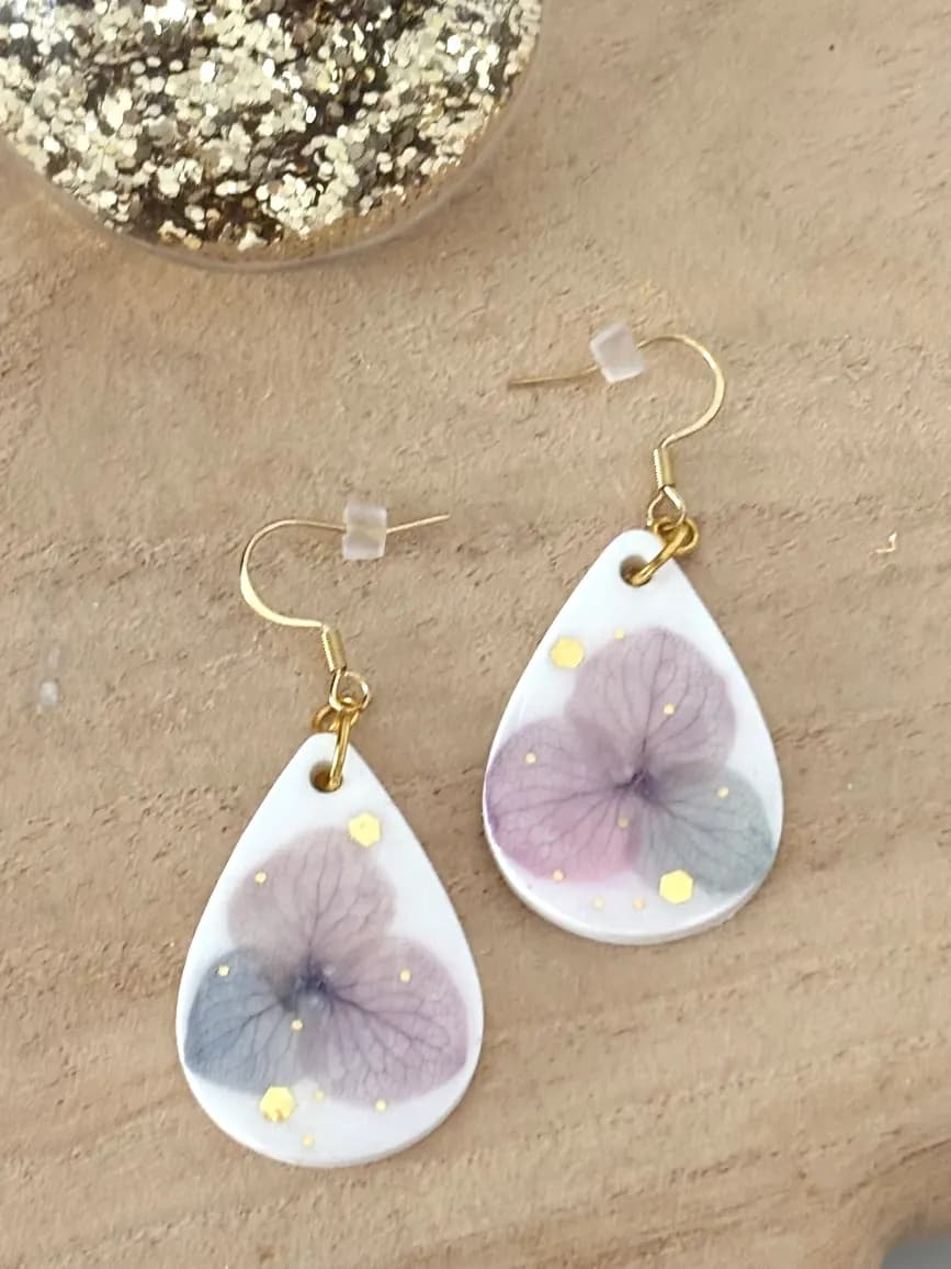 Boucles d'oreilles pendantes blanches en goutte avec fleur mauve séchée et touches dorées sur fond bois.