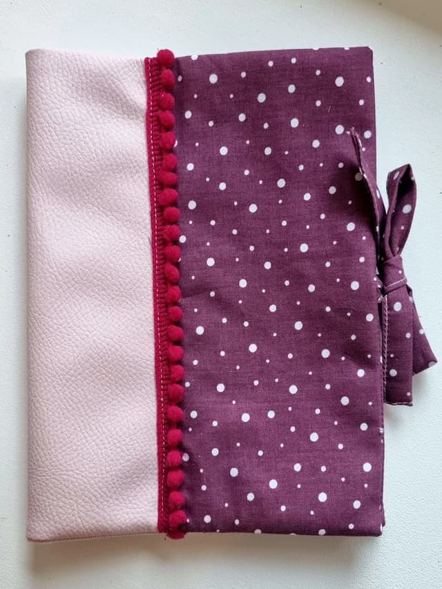 Protège-carnet de santé en simili cuir rose pâle et tissu prune à pois blancs, bordé de pompons et fermé par un lien