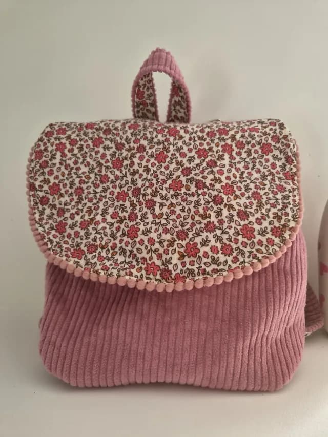 Vue de face du petit sac à dos vieux rose orné d'un rabat en tissu à fleurs et bordure de mini pompons.