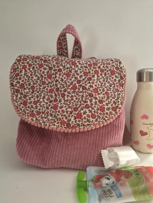Sac à dos pour enfant en velours côtelé rose avec rabat fleuri à pompons, posé près d'une gourde et d'un goûter.