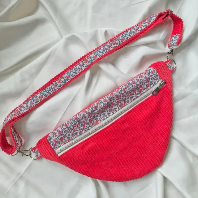 Sac banane rouge zippé avec sangle et détails à motifs floraux sur drap blanc plissé.