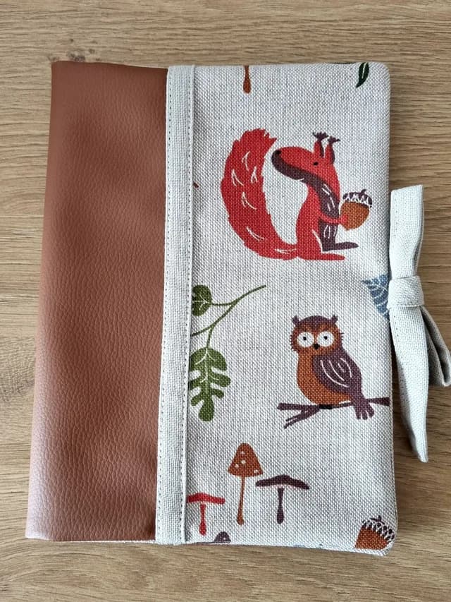 Protège-carnet fermé en simili cuir marron et tissu imprimé écureuil et chouette, noué avec un ruban.