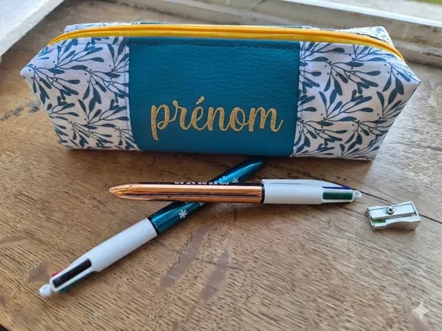 Trousse bleu canard et motif feuillage avec zip jaune et prénom doré, posée avec des stylos.