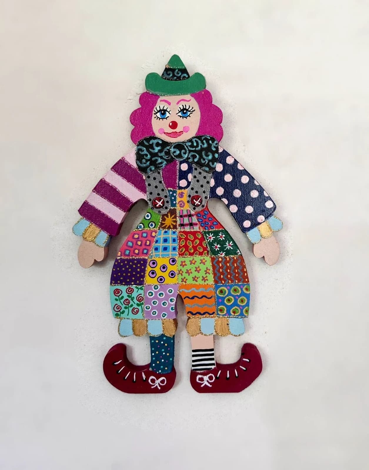 Figurine murale en bois d'un clown peint aux motifs multicolores façon patchwork avec chapeau vert et cheveux roses.